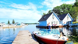 Een houten steiger die uitloopt in kalm water, met een rode boot afgemeerd aan het einde. Achtergrond toont een wit huis met een donker dak en een blauw raam.