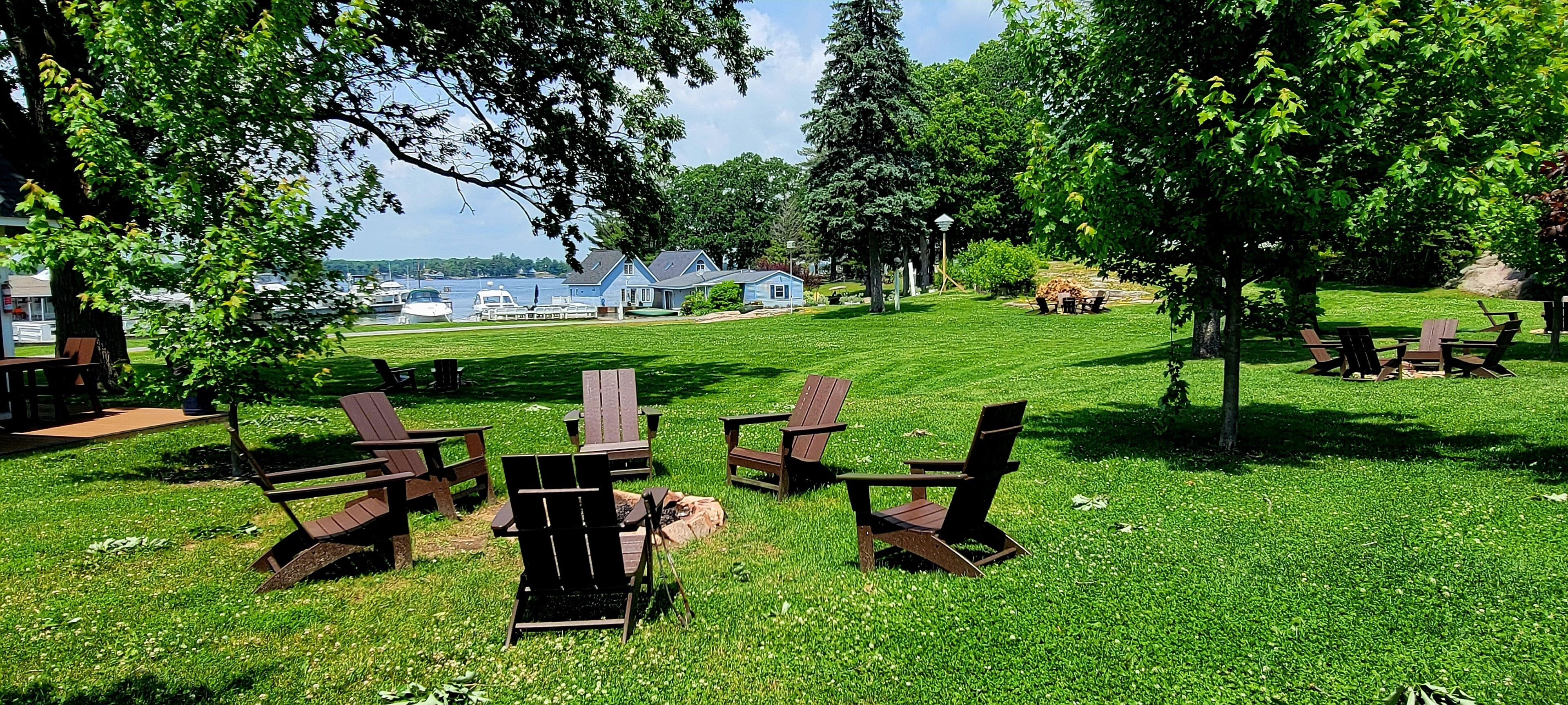 A grassy outdoor area with wooden Adirondack chairs and tables, a small building in the background, and a body of water.