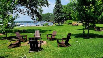 A grassy outdoor area with wooden Adirondack chairs and tables, a small building in the background, and a body of water.