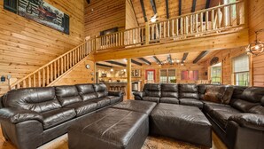 A spacious living area with a large L-shaped sectional sofa and ottoman, a wooden staircase, and a dining area in the background.