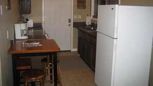 Cocina compacta con refrigerador, lavamanos y cafetera. Hay una mesa de madera con dos taburetes y una puerta que da a otra habitación.