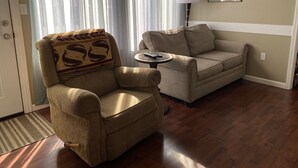 Una sala de estar con un sillón a rayas, un sofá, una mesita redonda con una lámpara y una ventana con cortinas traslúcidas.