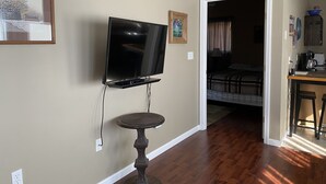 Una habitación con una televisión de pantalla plana en la pared, una mesita con una lámpara y una cama al fondo.