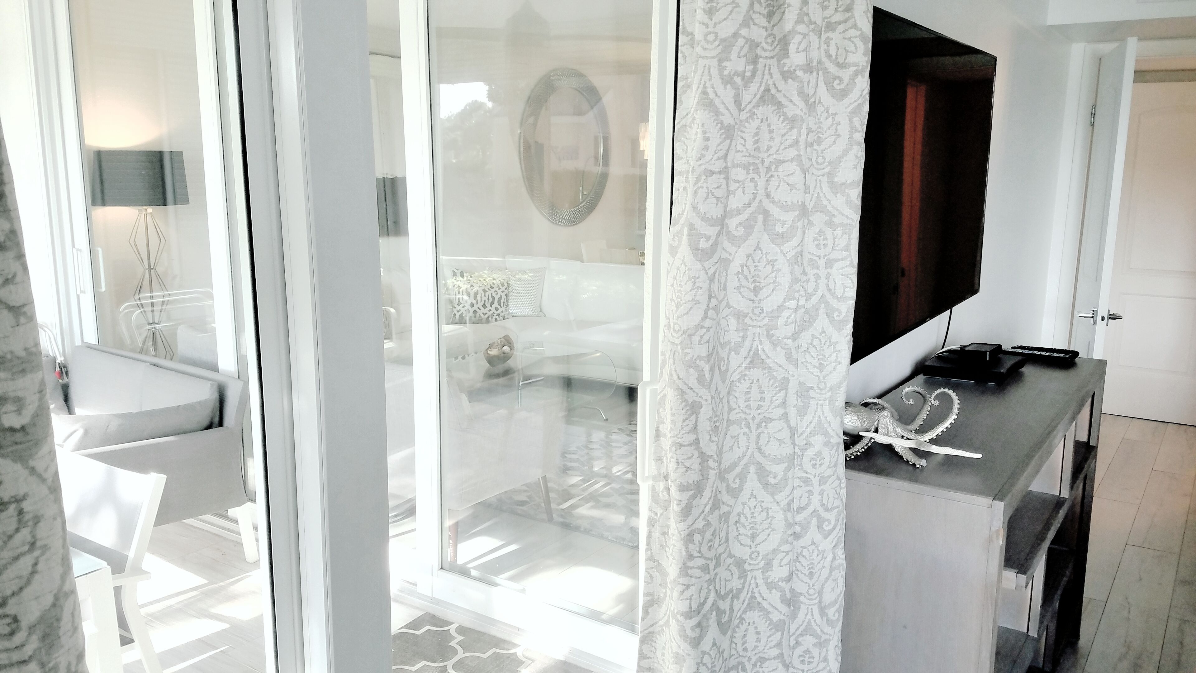 Una habitación de hotel moderna con un vidrio corredizo que da a un balcón, un sofá blanco, una mesita y un televisor sobre un mueble.