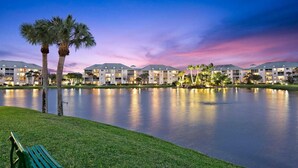 A hotel with a large body of water in front, palm trees, and a bench. - POSHPADZ-Bella Lago-Ground Floor, Walk to Jupiter Beach, Patio with Lake Views (Jupiter)
