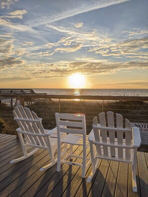 Twee Adirondack-stoelen op een houten terras met uitzicht op een strand bij zonsondergang.