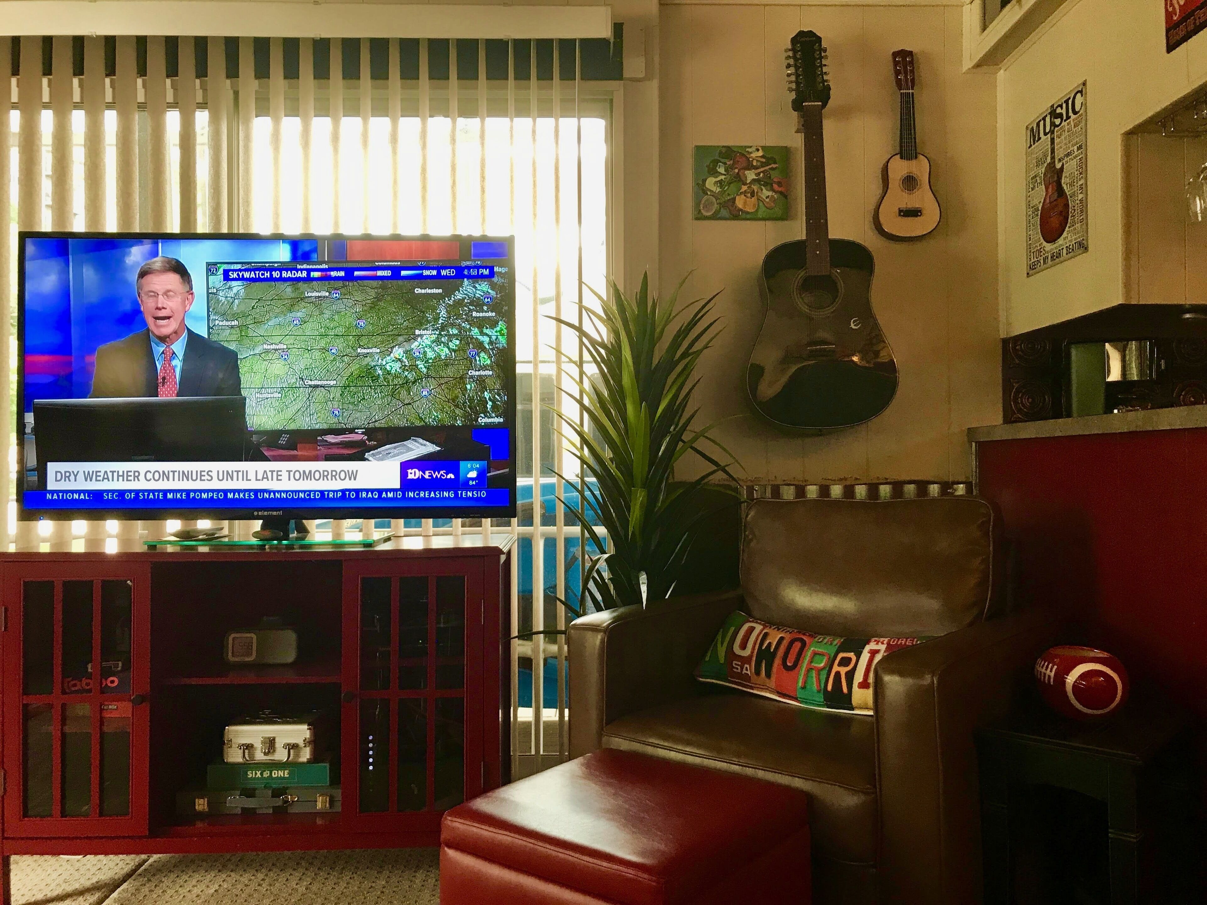 Una sala de estar con una televisión mostrando un noticiero, una guitarra y un sillón de cuero.