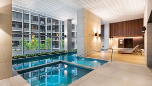 Una piscina interior con paredes de vidrio transparente, un área de descanso con tumbonas y vistas a un edificio con balcones.