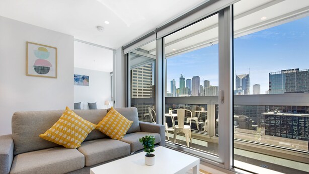 Una sala de estar moderna con un sofá gris, cojines estampados amarillos, una mesa de centro blanca y vistas a la ciudad a través de amplios ventanales.