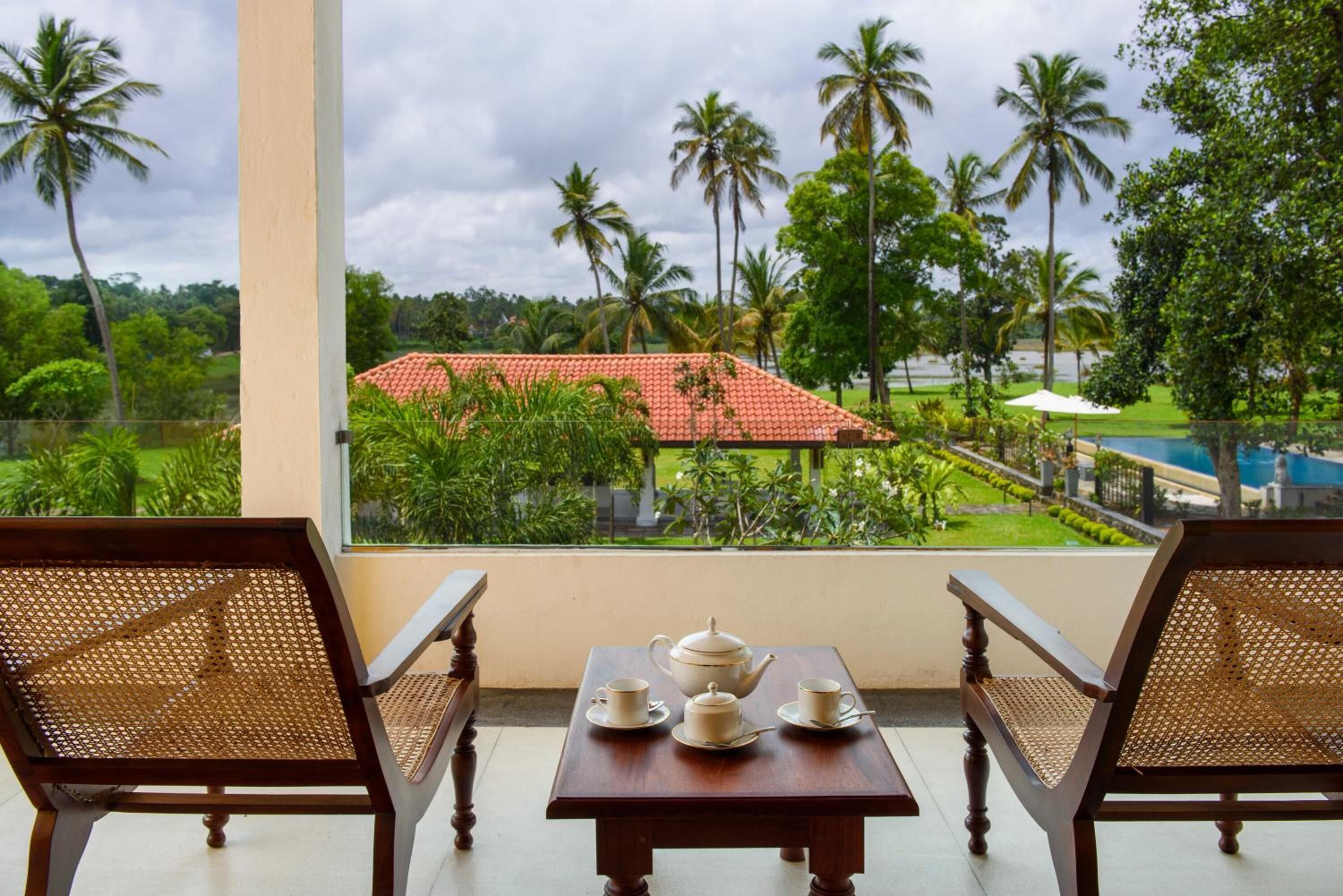 A balcony with two wicker chairs, a small table with a teapot and cups, and a view of a red-roofed building surrounded by palm trees.