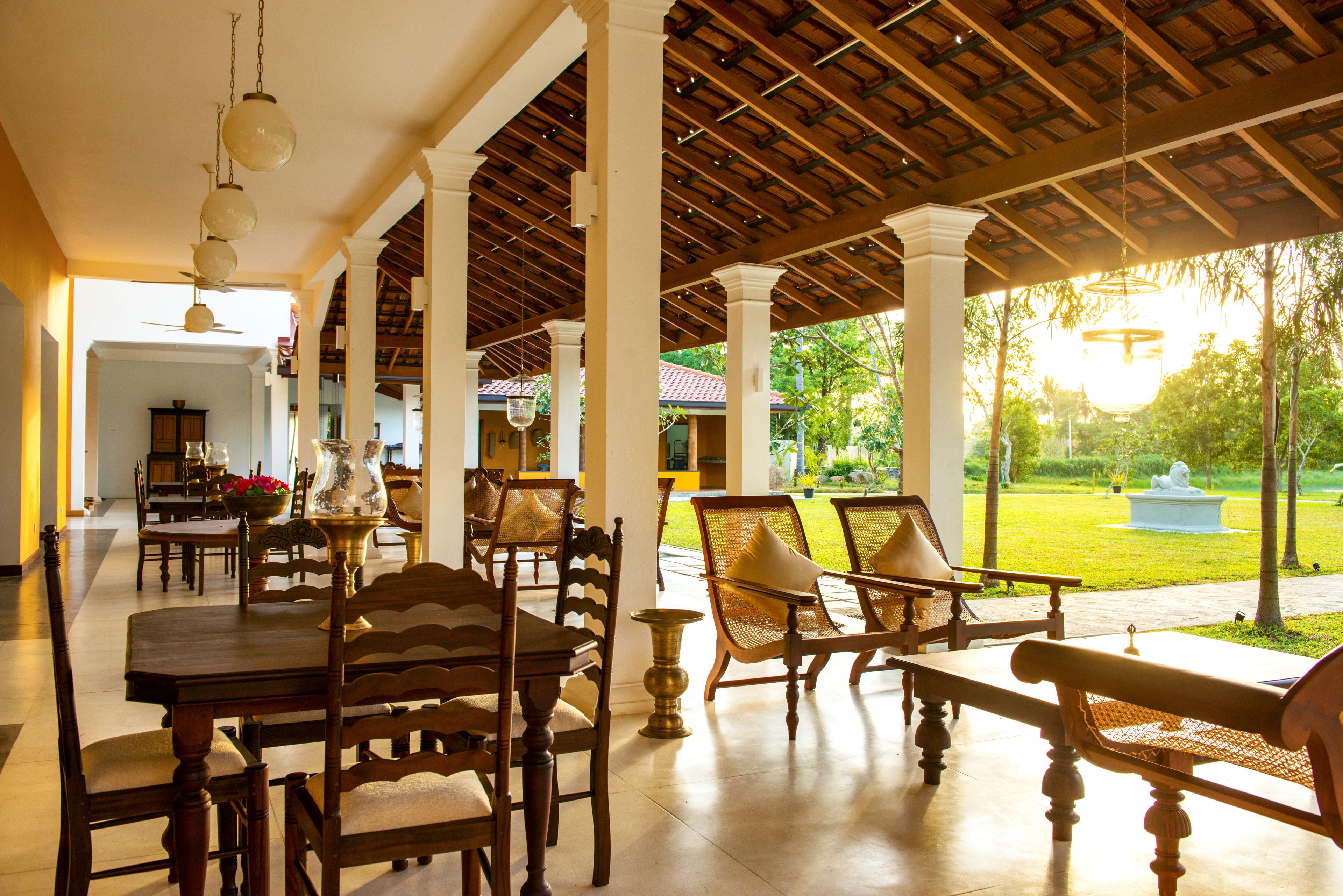 A covered outdoor dining area with wooden tables and chairs, a ceiling with wooden beams, and a view of a garden with a statue.