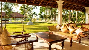 A covered outdoor seating area with wicker furniture and a table, overlooking a well-maintained lawn and palm trees. - The Notary's House (Pannala)