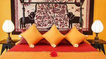 A bed with three orange pillows, a red bedspread, and a decorative tapestry with birds and floral patterns on the wall behind.
