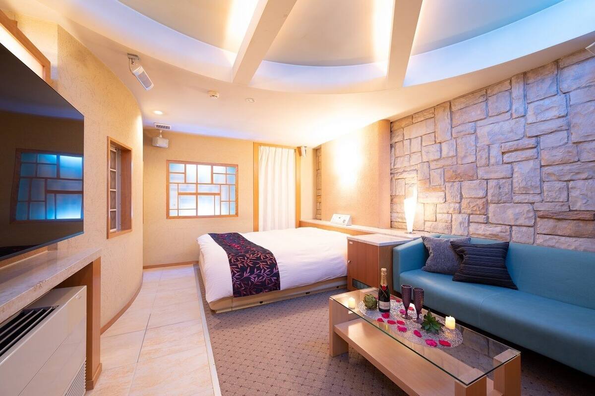 A modern hotel room with a stone accent wall, a bed, a sofa, a TV, and a glass coffee table with a bottle and candles.