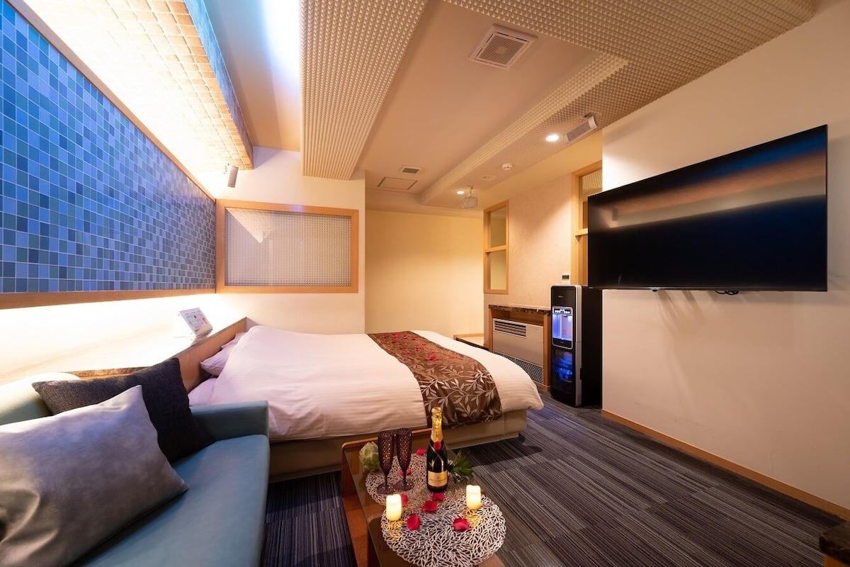 A modern hotel room with a large bed, a flat-screen TV, a seating area with a small table, and a large window with blue-tiled walls.