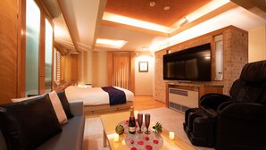 Habitación de hotel con cama, sofá, sillón de masaje, televisión y una mesita con velas y una botella.