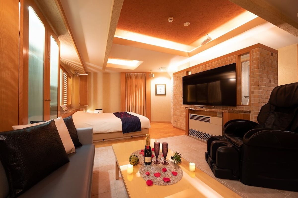 A hotel room with a bed, a sofa, a massage chair, a TV, and a small table with candles and a bottle.
