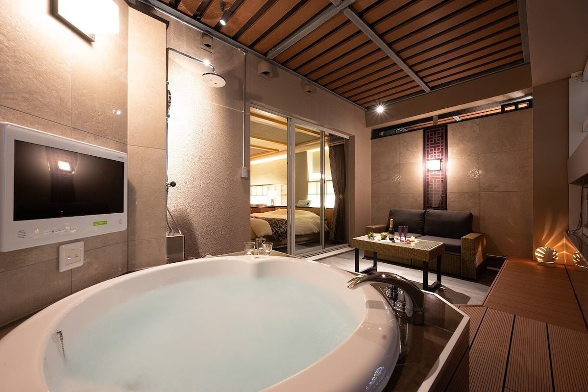 A modern bathroom with a large bathtub, a television, and a sliding glass door leading to a bedroom.