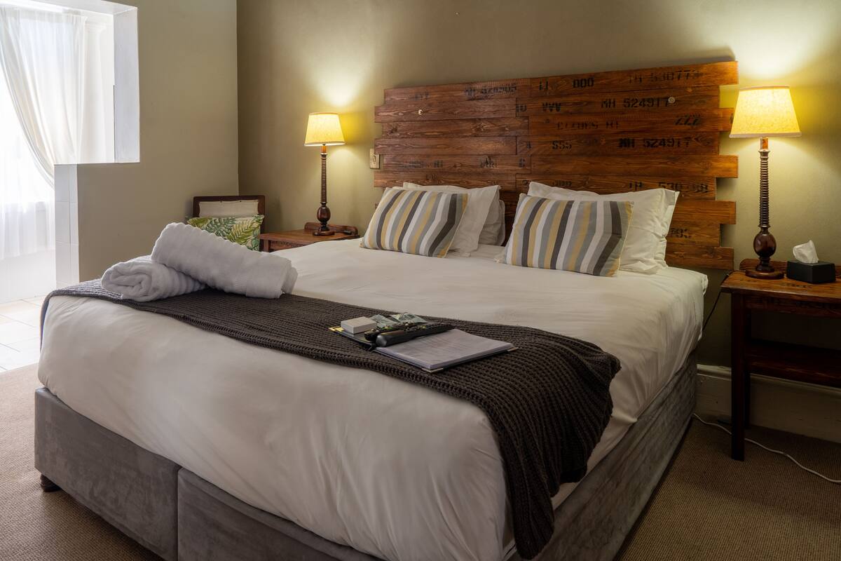 Una cama bien hecha con un almohadón a rayas, un control remoto y un libro sobre la cama. Hay dos lámparas de noche sobre mesitas de madera.