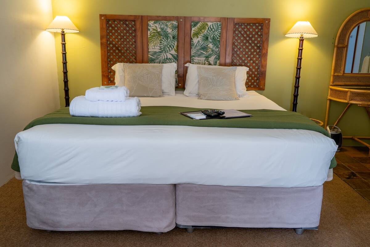 Una cama doble con una colcha verde, dos almohadas blancas y un juego de toallas sobre la cama. Hay dos lámparas fijadas a la pared a cada lado de la cama.