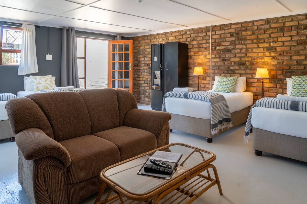 Una habitación de hotel con un sofá marrón, dos camas individuales, un armario y una mesa de centro de vidrio.