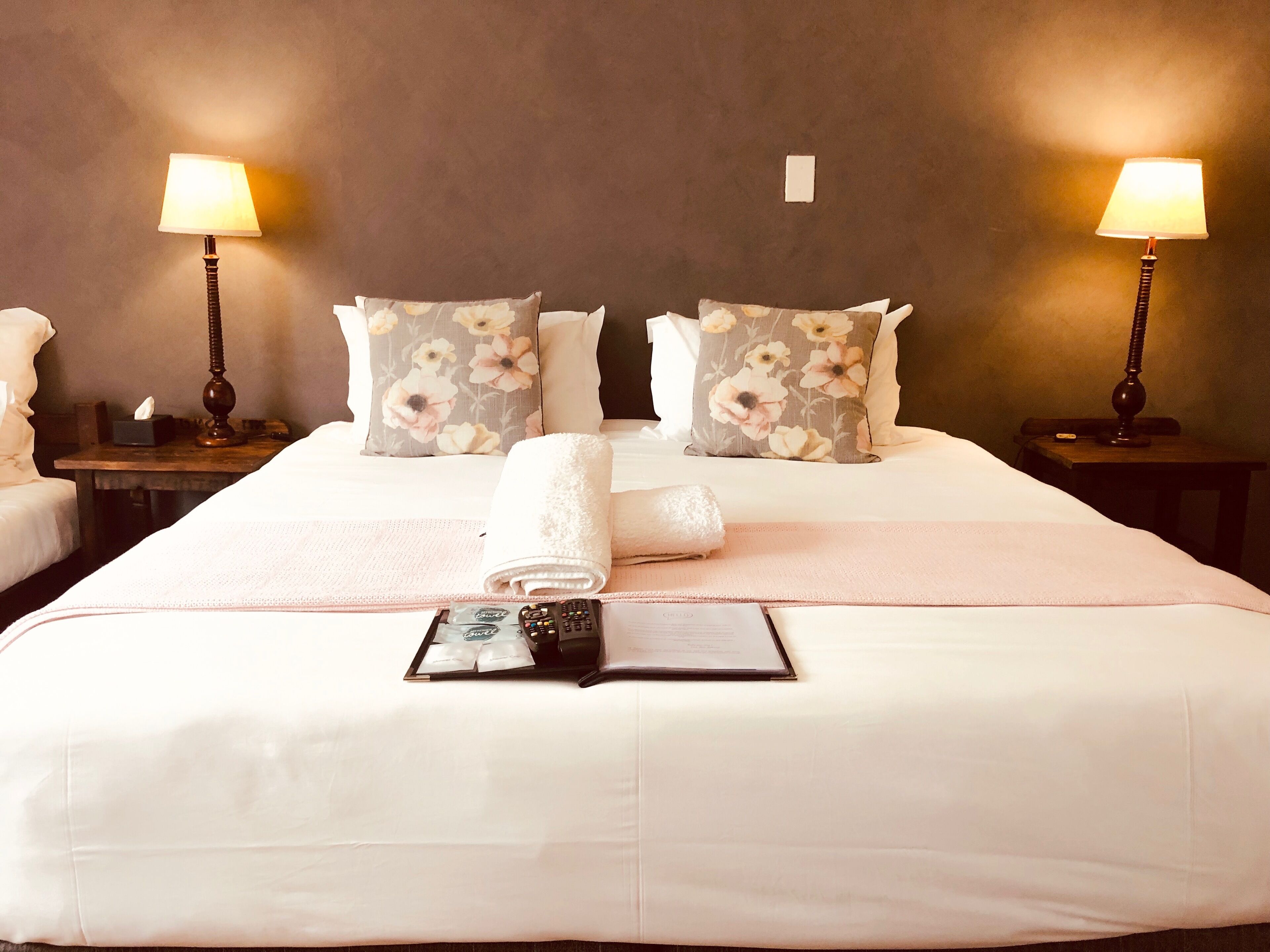 A hotel room with a large bed, two bedside tables, a lamp on each table, floral-patterned pillows, a rolled white towel, and a remote control on the bed.