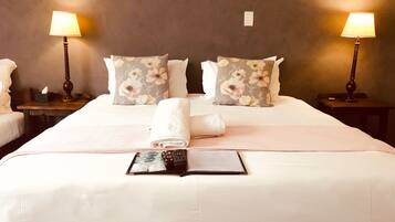 A hotel room with a large bed, two bedside tables, a lamp on each table, floral-patterned pillows, a rolled white towel, and a remote control on the bed.