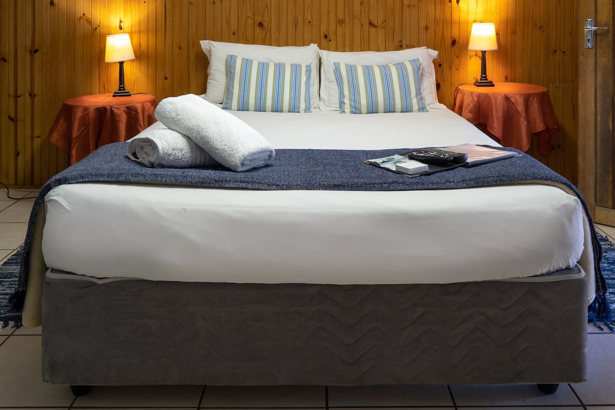 Una cama bien hecha con almohadas blancas y a rayas, una toalla doblada y un control remoto sobre la cama. Hay dos mesitas redondas con lámparas a cada lado de la cama.