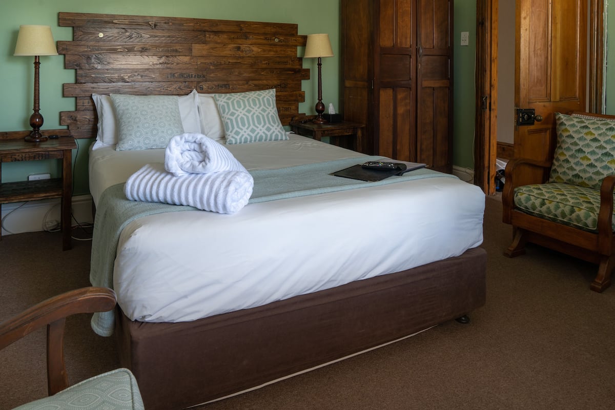 Una cama bien tendida con cabecera de madera, una mesita de noche con lámpara y una silla con cojín estampado.