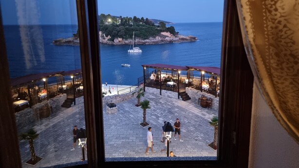 Une vue depuis une fenêtre donnant sur une zone côtière, avec une terrasse où l’on peut prendre ses repas, une petite île et une étendue d’eau.