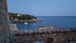 Un balcon avec des fauteuils en osier et une table en verre, donnant sur une zone côtière où l’on aperçoit des bateaux et un bâtiment situé sur une colline.