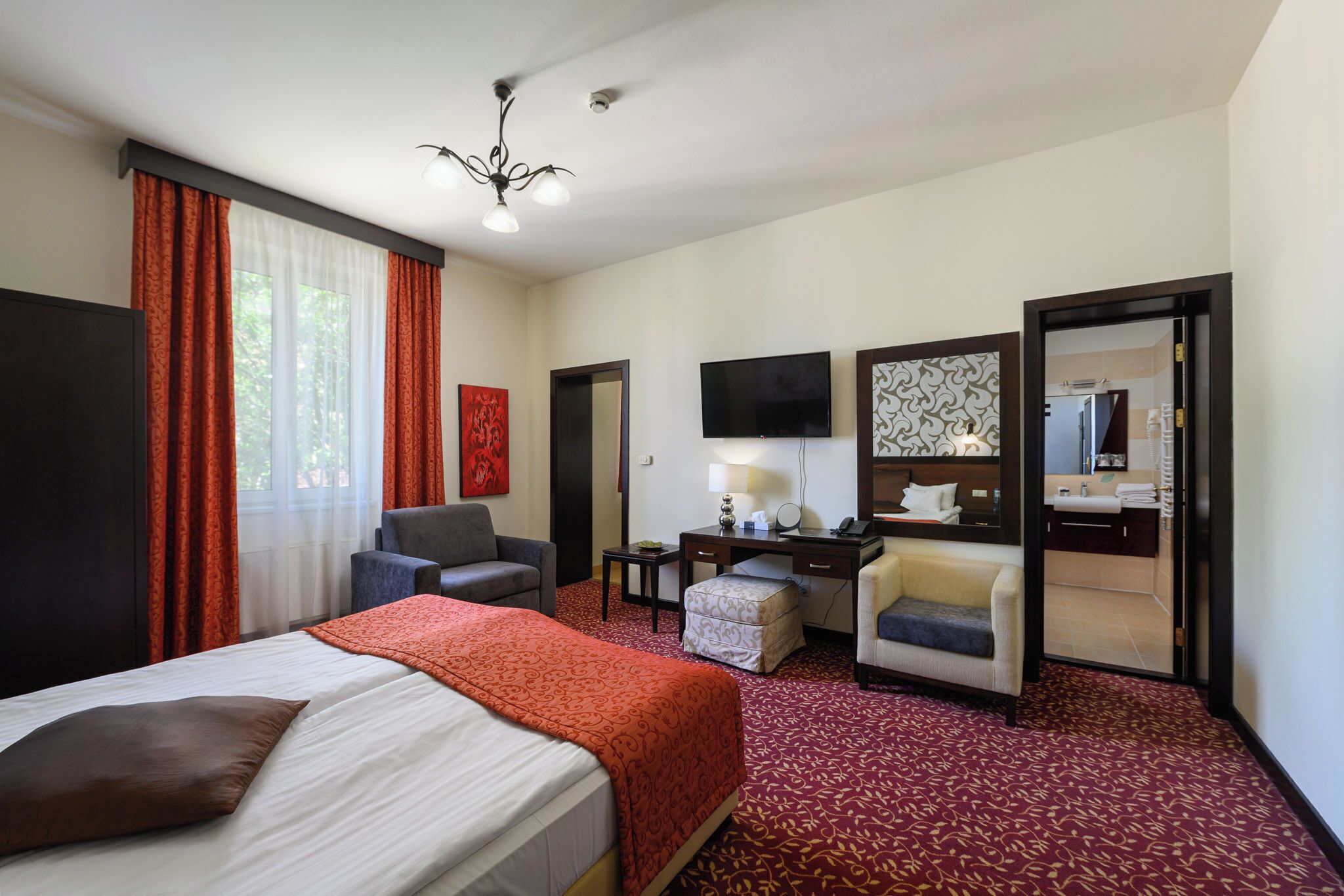 A hotel room with a bed, two armchairs, a desk with a lamp, a television, and a bathroom visible through an open door.