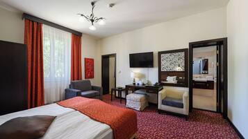 A hotel room with a bed, two armchairs, a desk with a lamp, a television, and a bathroom visible through an open door.