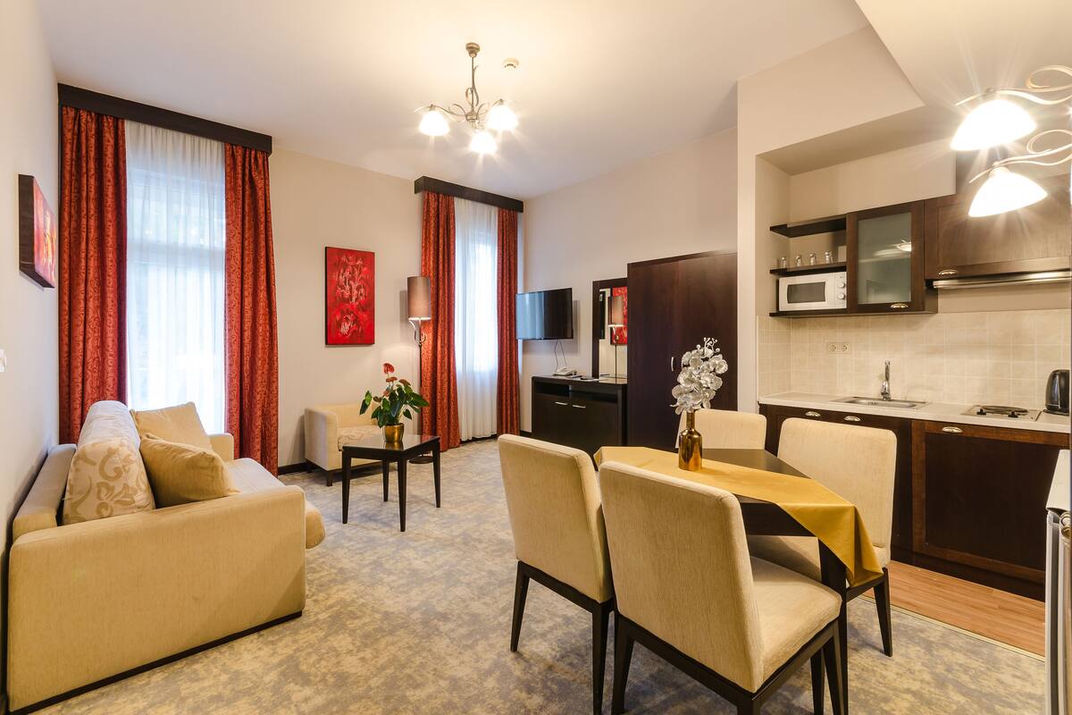 A modern living room with a sofa, dining table, and chairs, a kitchen with a microwave and cabinets, and a TV on the wall.