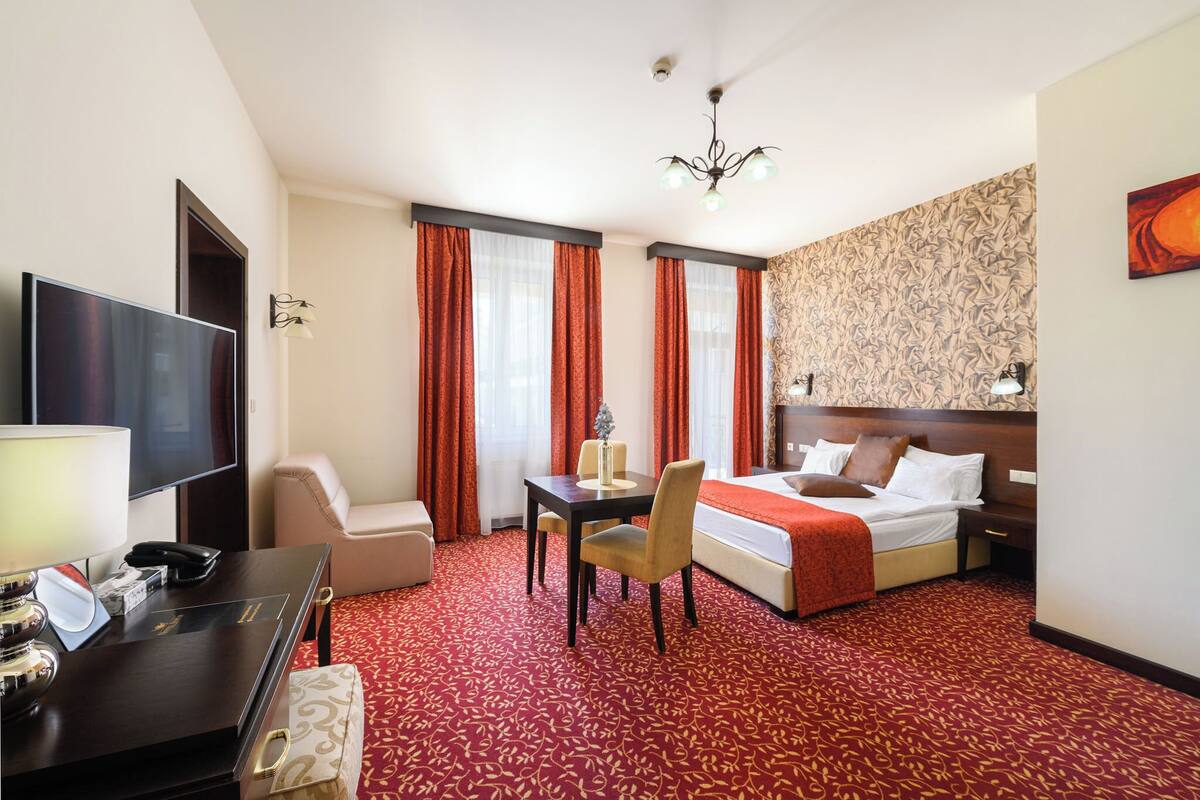 A hotel room with a bed, a desk with a telephone, a television, and a red carpeted floor.