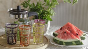 Une théière en verre, deux verres et une assiette avec des tranches de pastèque, le tout posé sur un plateau en bois.