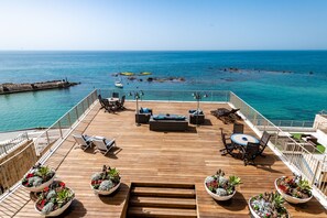 Een houten terras met buitenmeubels, bloempotten en uitzicht op zee.