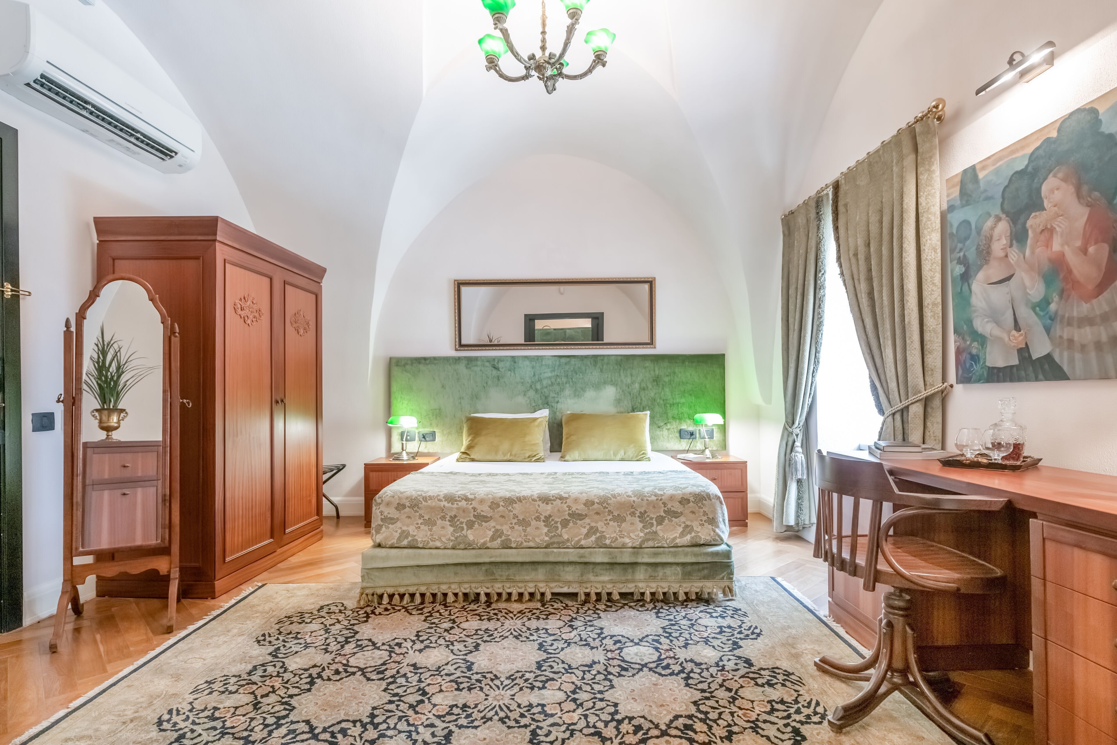 Ein Schlafzimmer mit einem großen Bett, einem Schreibtisch, einem Stuhl, einem Kleiderschrank, einer Chandelierleuchtre und einem Gemälde an der Wand.