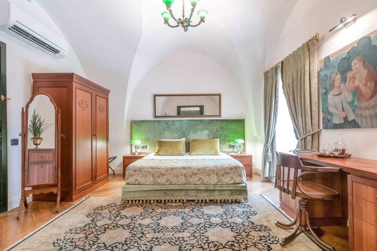 Ein Schlafzimmer mit einem großen Bett, einem Schreibtisch, einem Stuhl, einem Kleiderschrank, einer Chandelierleuchtre und einem Gemälde an der Wand.