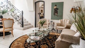 A spacious living room with a round glass coffee table, a beige sofa, a white armchair, and a staircase with ornate black railings.