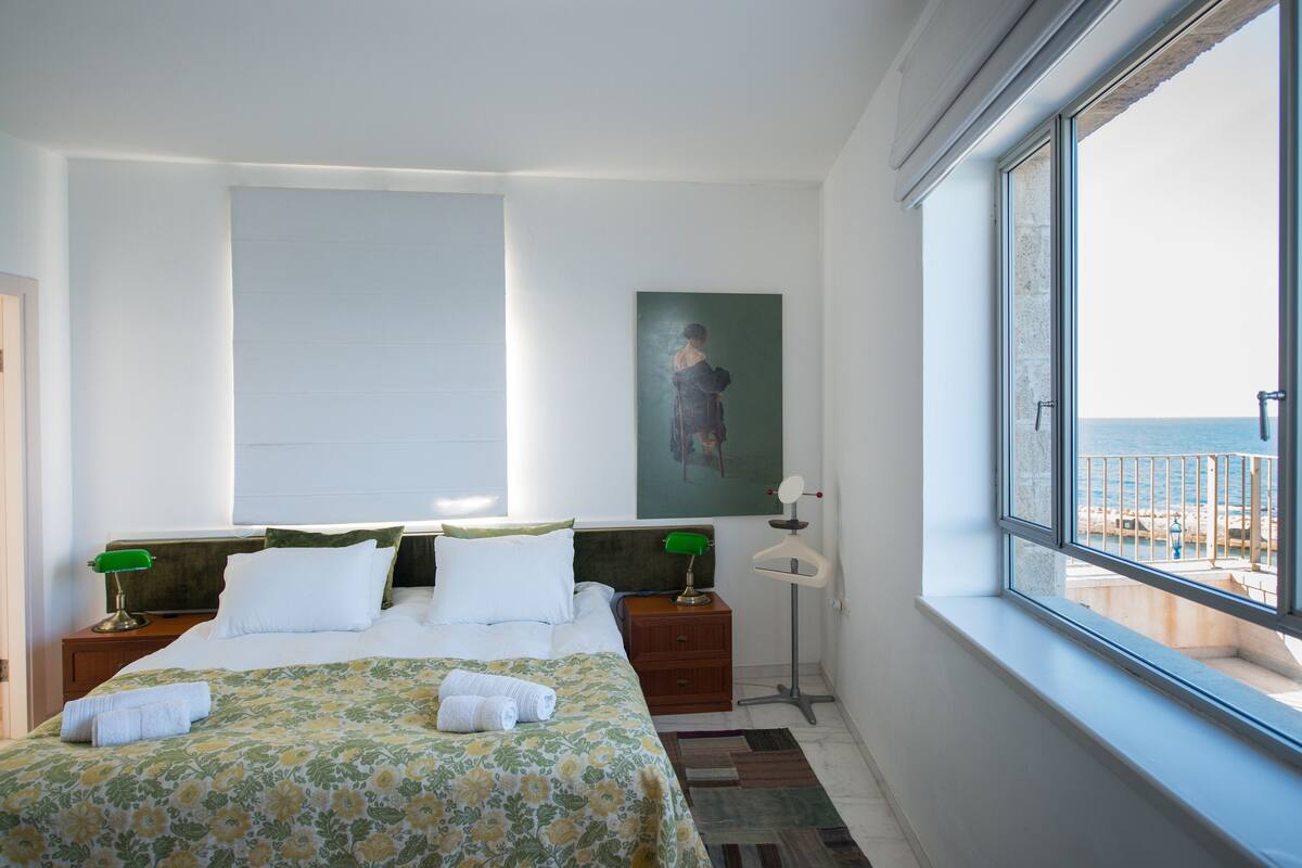 Ein Hotelzimmer mit Bett, Nachttisch, Lampe, einem Gemälde und Meerblick.