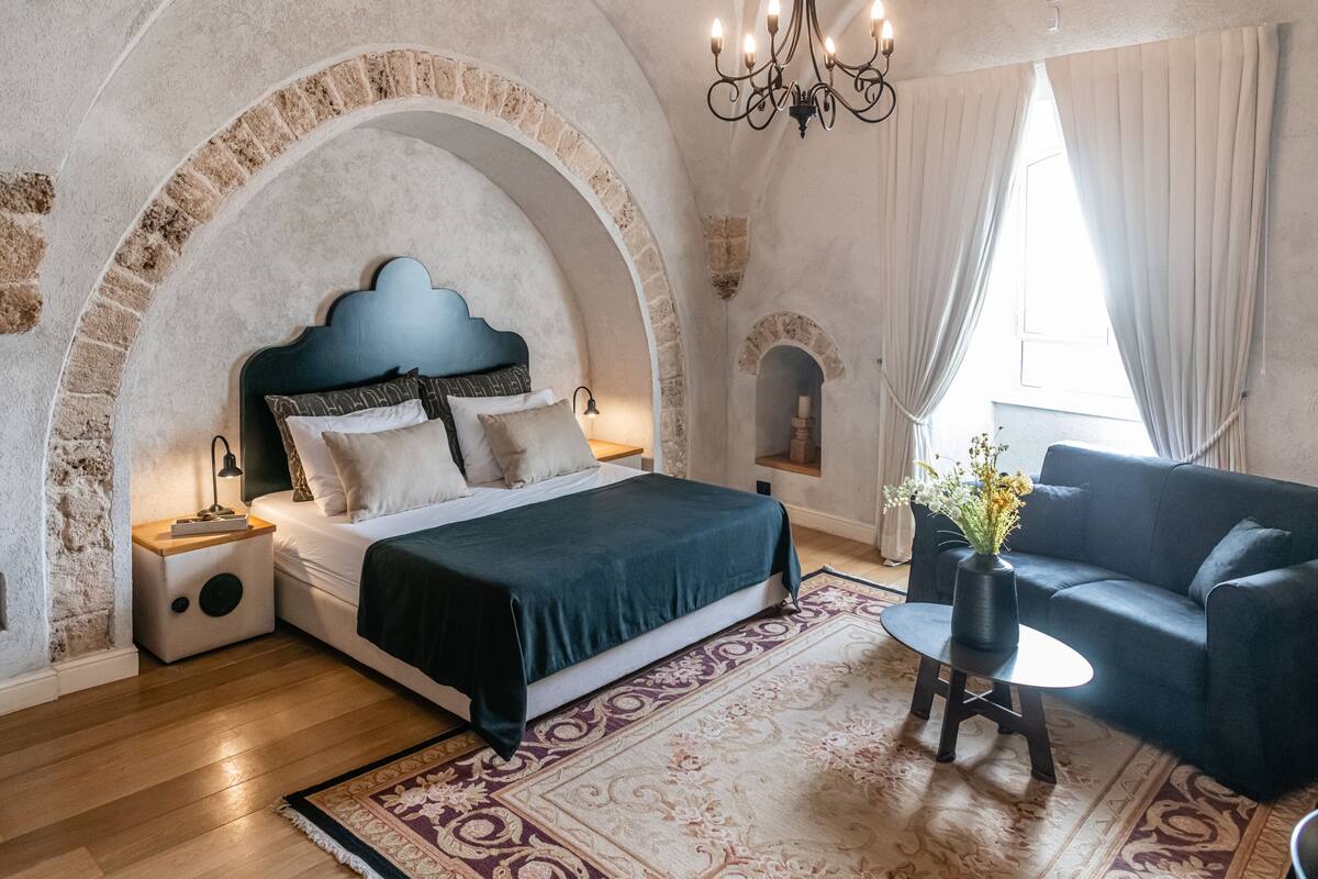 Ein Schlafzimmer mit einem Bett, einem Nachttisch, einer Couch, einem kleinen Tisch mit einer Vase und einer gewölbten Steinwand.