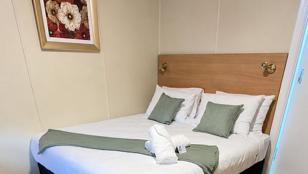 Un lit bien fait, avec du linge de lit blanc et des touches de vert, une tête de lit en bois et une œuvre d’art florale encadrée accrochée au mur.