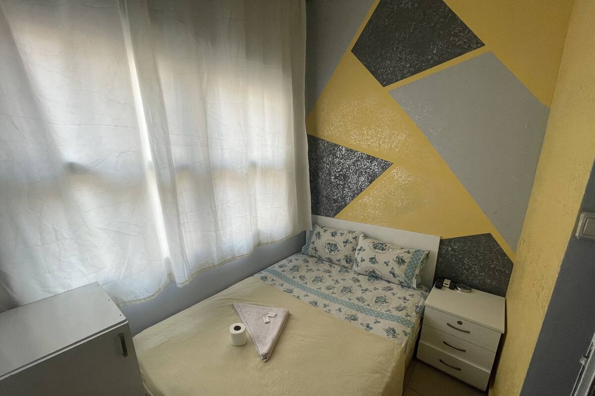 A small, neatly arranged bedroom with a bed, a nightstand, a drawer unit, and a curtain.
