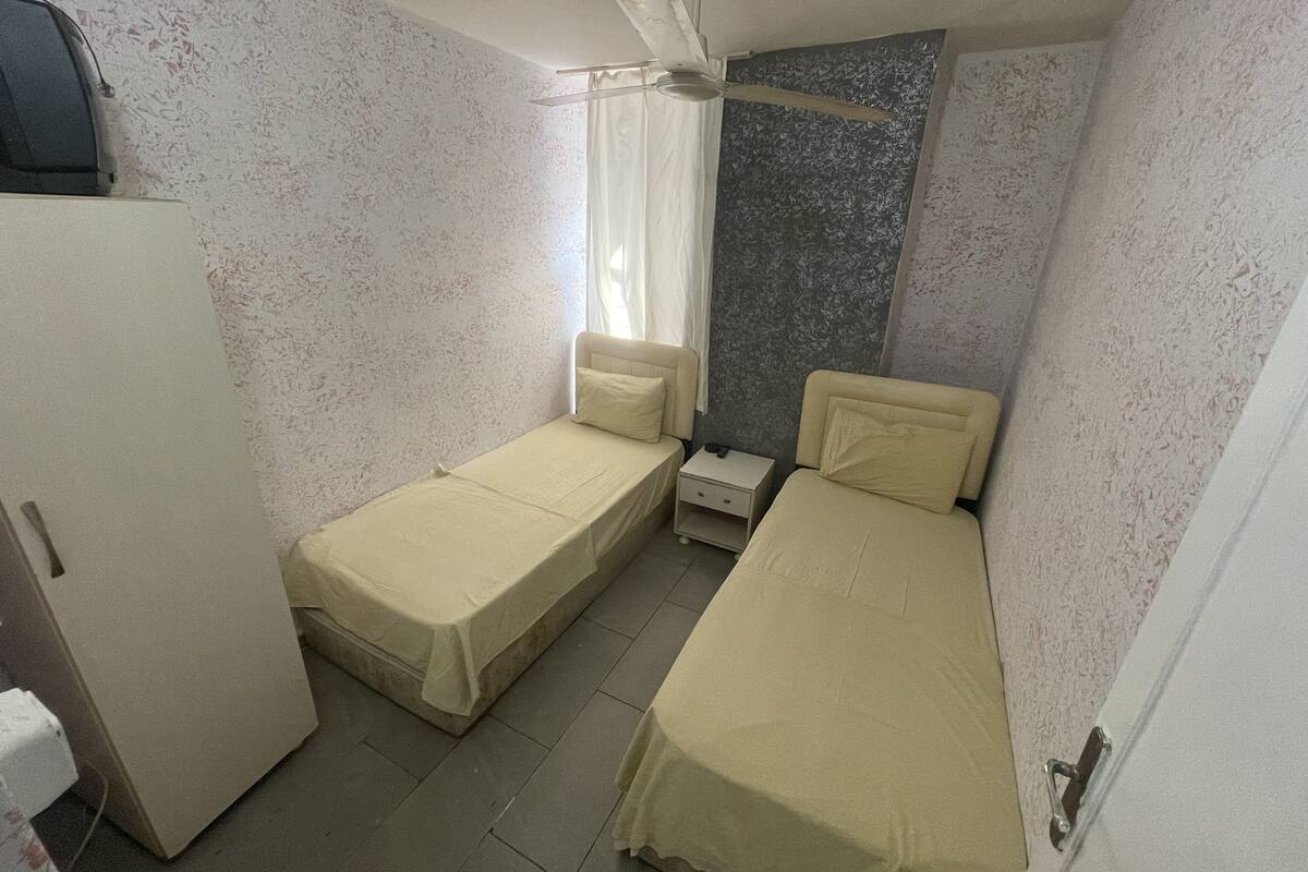A small, compact room with two single beds, a small bedside table, a wardrobe, and a ceiling fan.