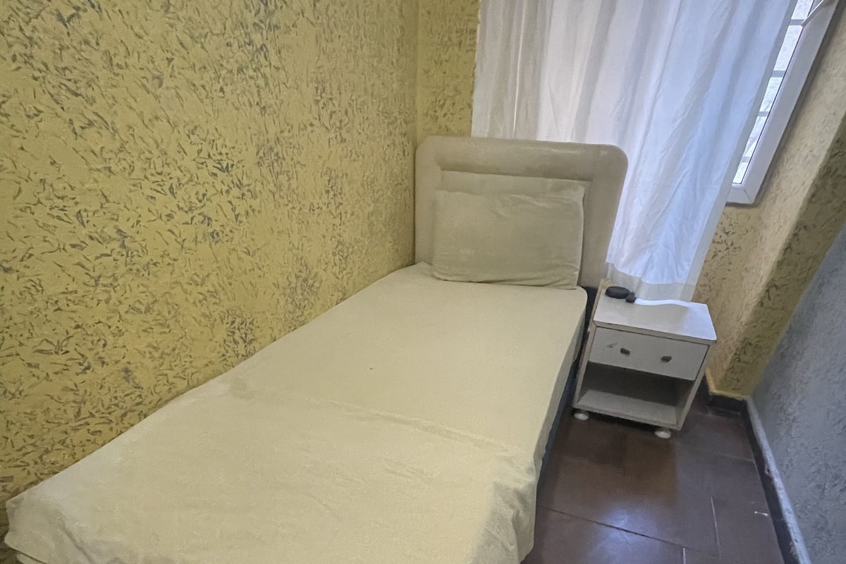 A small, single-bed room with a plain bed, a bedside table, and a nightstand. The walls have a textured pattern, and there's a window with a white curtain.