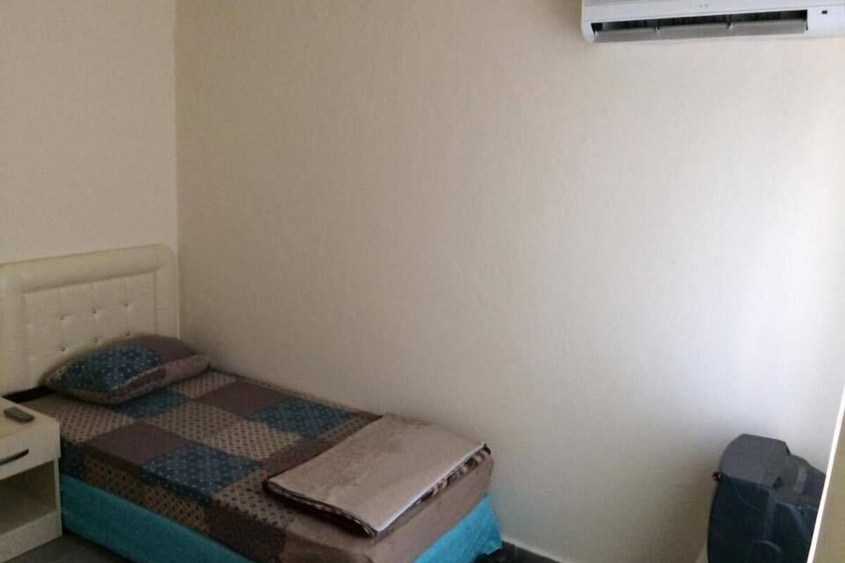 A small, simple room with a single bed, a nightstand, a wall-mounted air conditioner, and a trash bin.