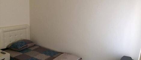 A small, simple room with a single bed, a nightstand, a wall-mounted air conditioner, and a trash bin.