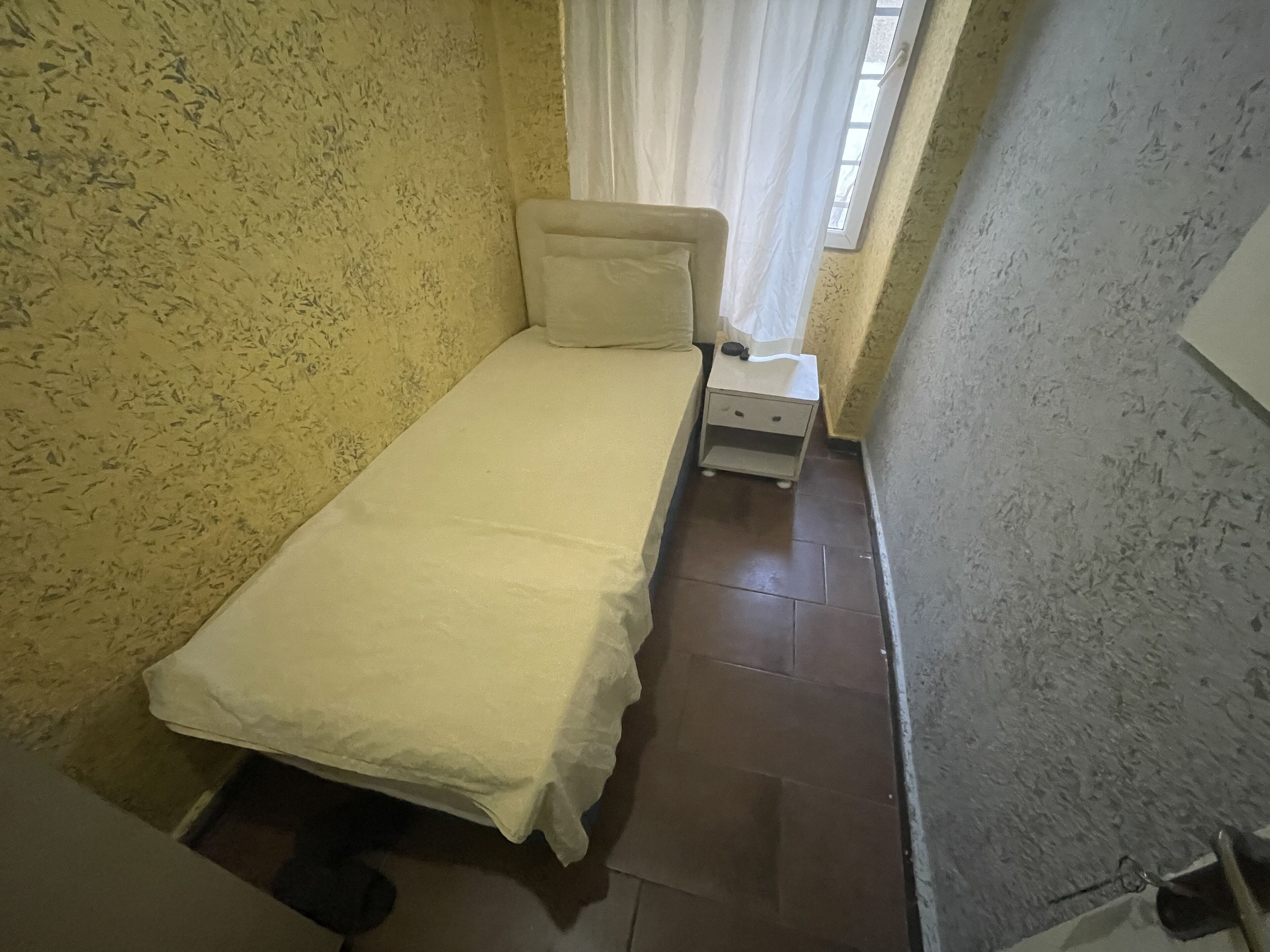 Ein kleiner, schmaler Raum mit einem Einzelbett, einem Nachttisch und einem fenster mit Vorhängen.