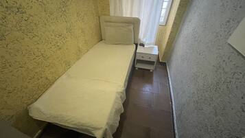 Ein kleiner, schmaler Raum mit einem Einzelbett, einem Nachttisch und einem fenster mit Vorhängen.